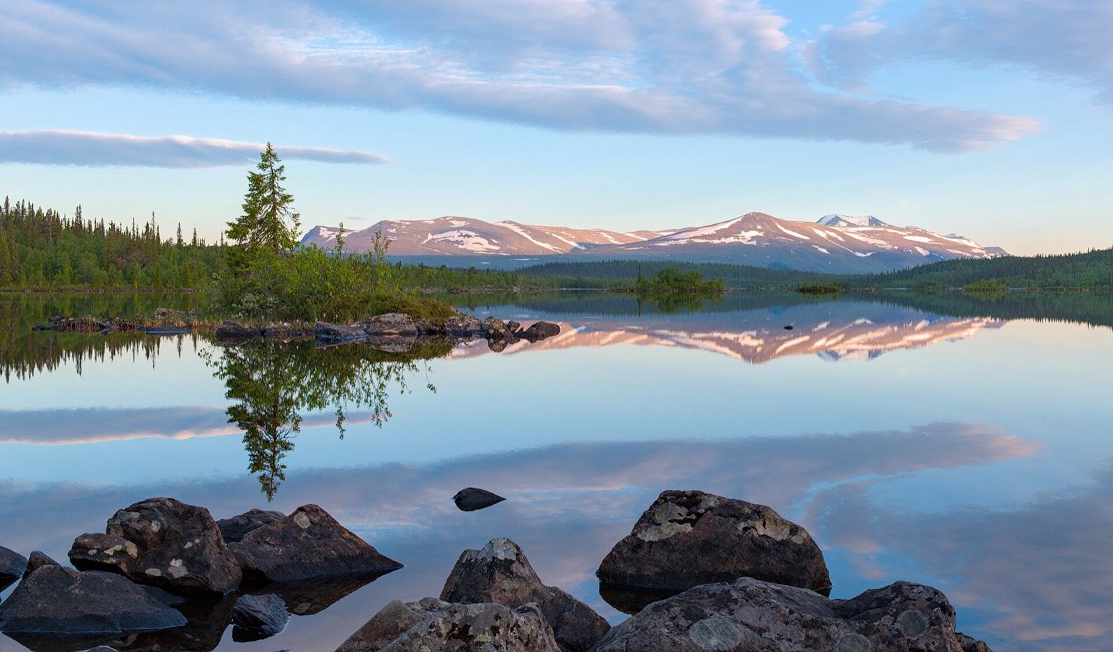 A still mountain lake at golden hour on the Kungsleden trail, snow patched fells reflected in the water and a lone spruce on a rocky islet