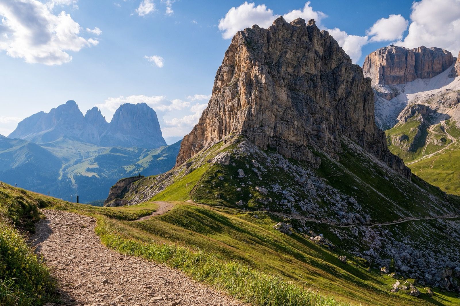 A gravel hiking trail curving through green alpine meadows toward the rocky towers of the Sella group in the Dolomites under a blue sky with white clouds