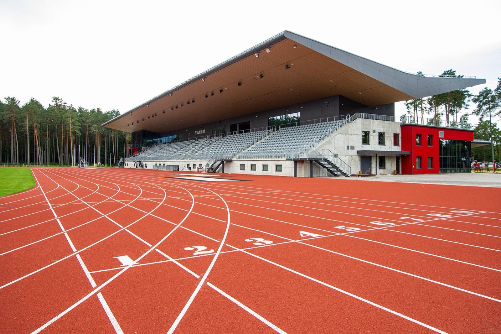 An empty red athletics track with white lane lines and numbered lanes curving in front of a modern grandstand at a stadium in Valmiera, Latvia